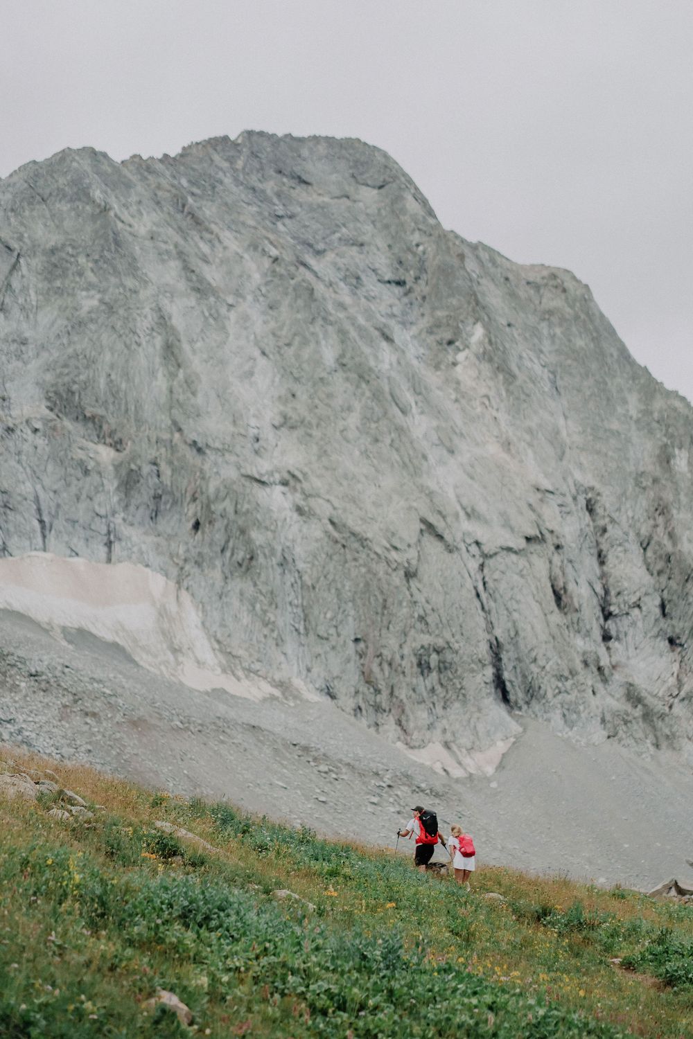 Tiny hikers trek beneath massive gray mountain face in alpine environment.