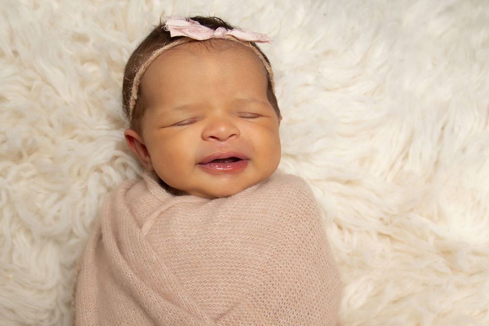 Newborn swaddled in beige wrap with pink bow smiles during professional photo session.
