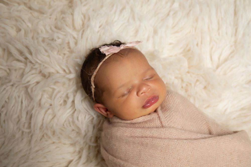 Newborn wrapped in beige swaddle rests on fluffy white fur backdrop.