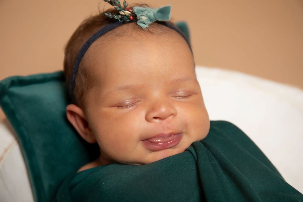 Sleeping newborn baby wearing green outfit and matching headband smiles contentedly.