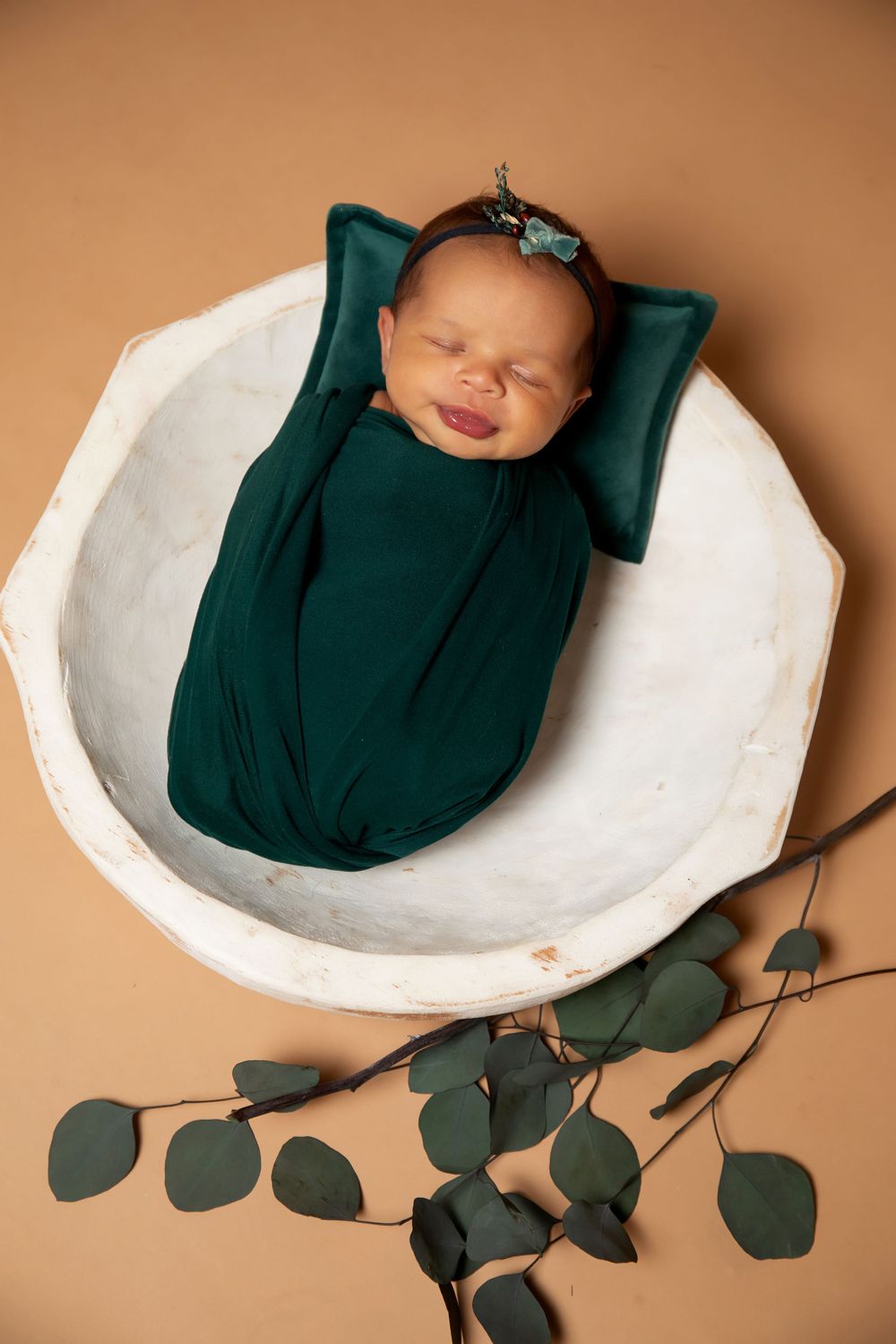 Sleeping newborn in emerald wrap posed in white bowl with eucalyptus decoration.