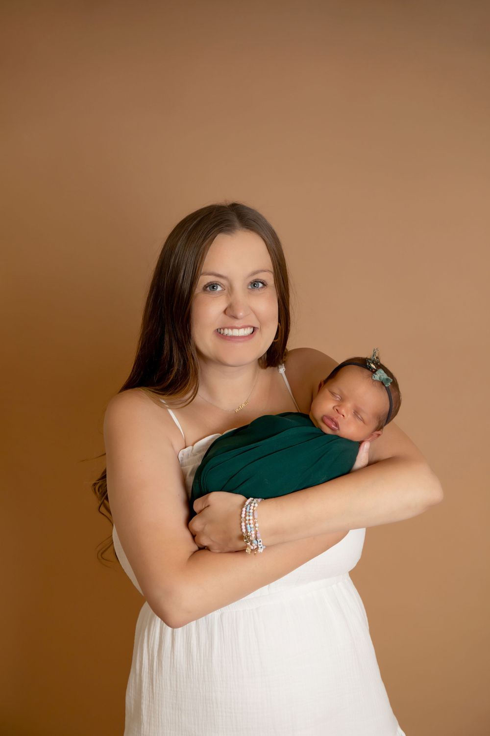 Mother in white dress holding swaddled newborn in emerald green against brown background.