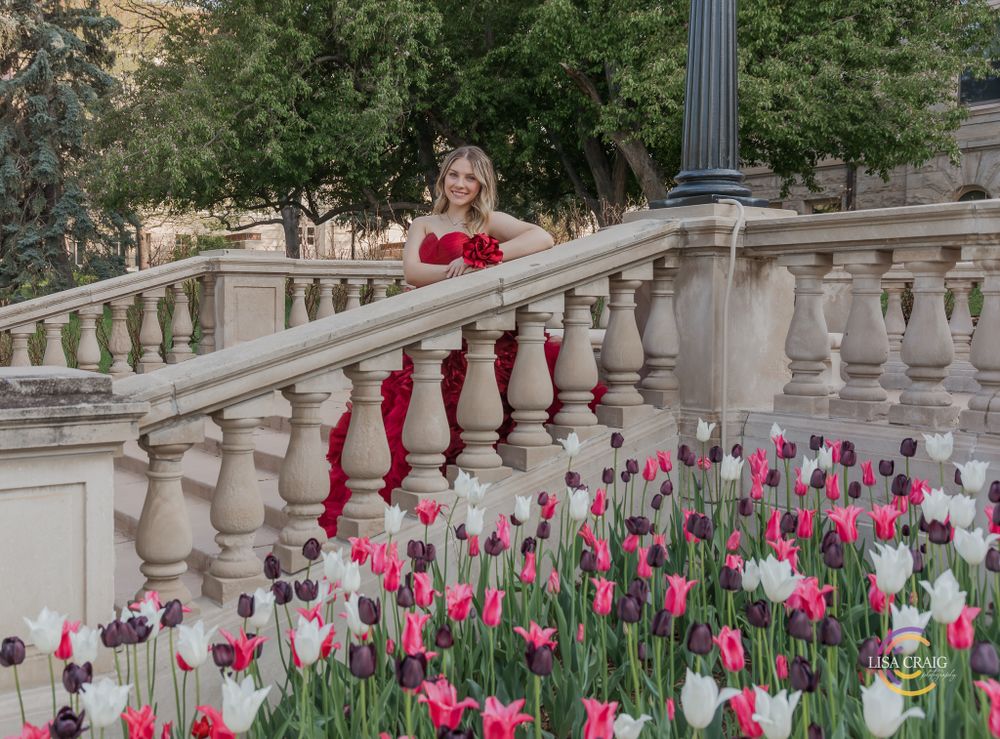 Person in red formal dress poses on stone balcony surrounded by pink and white tulips. Senior Portrait Photographer | Denver, Broomfield,
