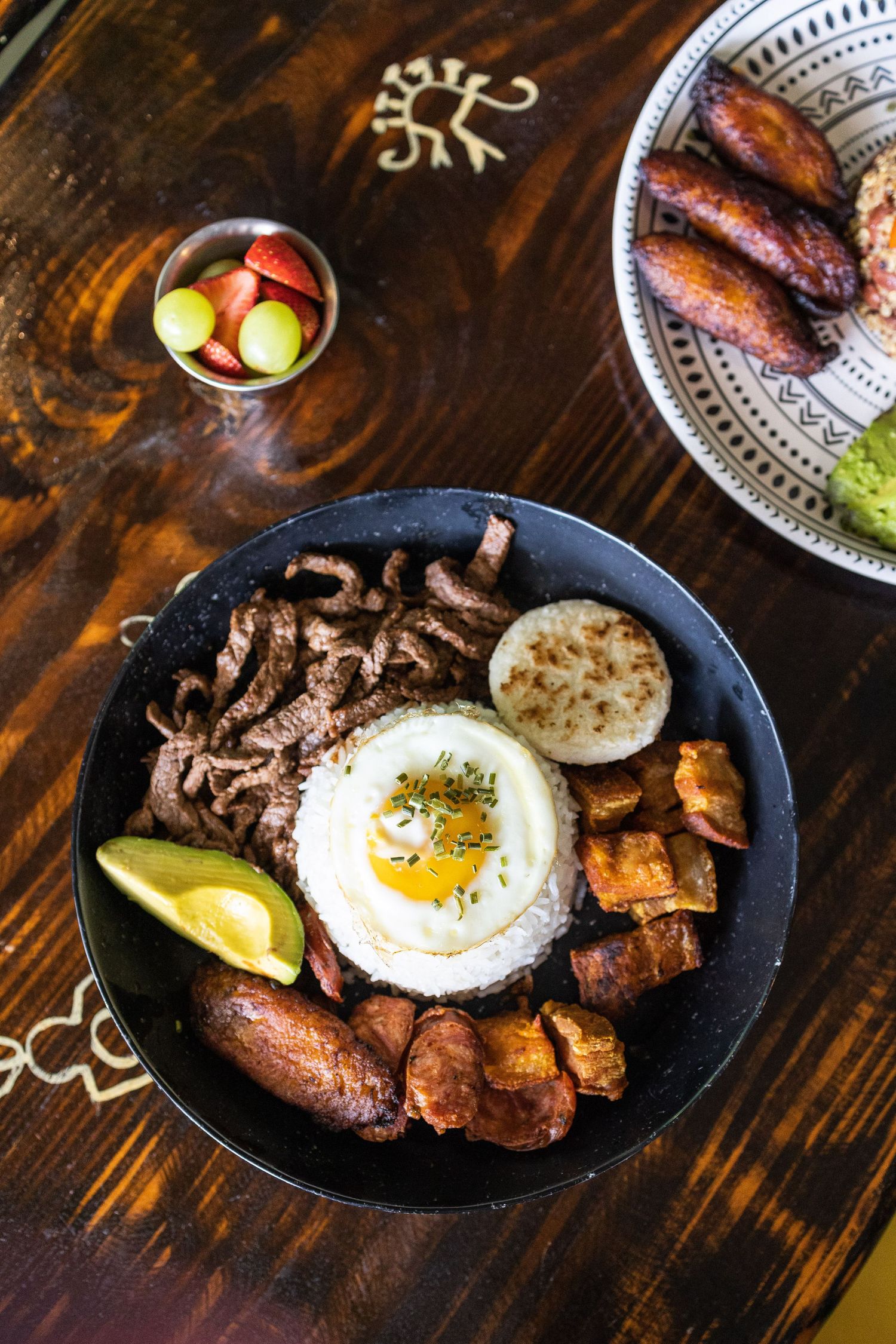 Rice bowl topped with sunny side up egg, beef, sausage and plantains on dark wooden table.