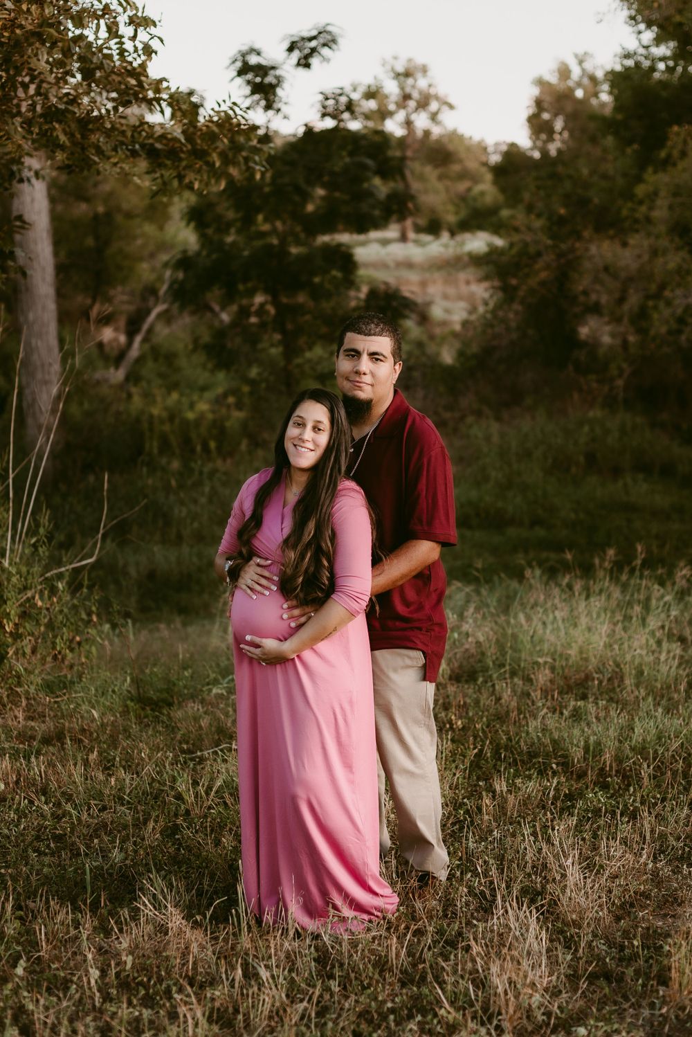 Couple stands together in a natural outdoor setting, wearing burgundy and pink.