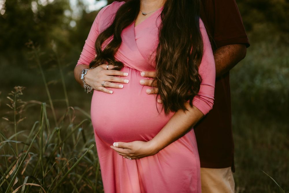 Detailed shot of expectant parents' hands resting on baby bump in pink maternity dress.