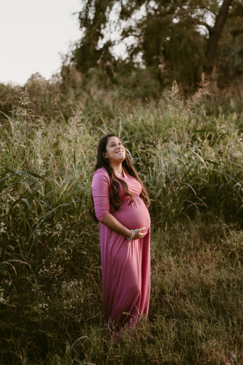 Maternity portrait showcasing woman in pink dress standing amid natural grassland setting.