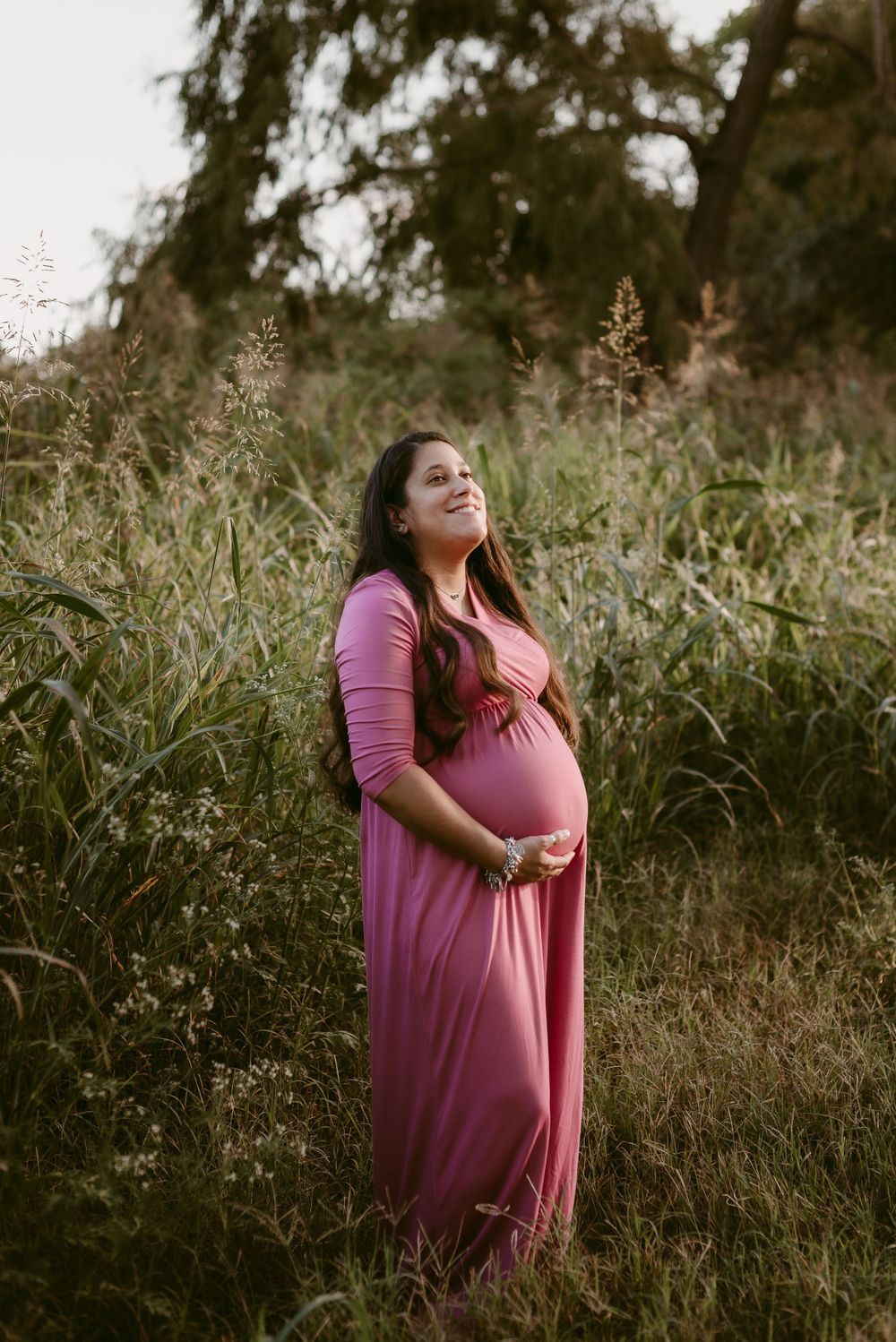 Woman in pink maxi dress cradles baby bump while standing among tall grass at sunset.