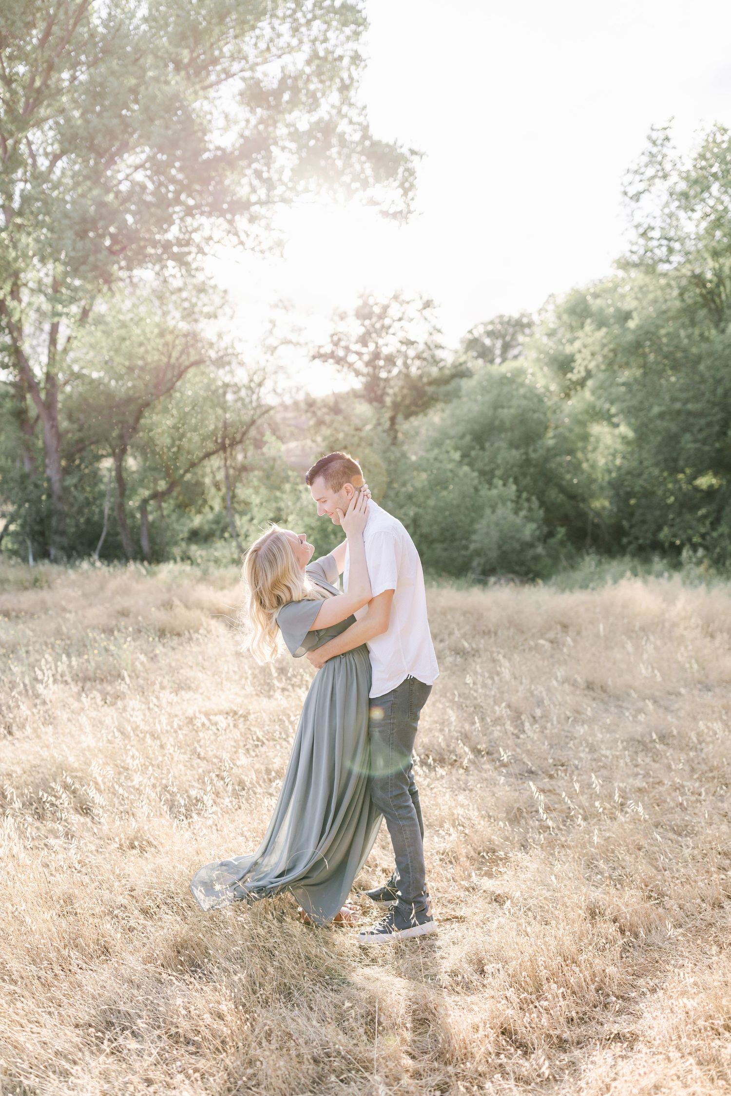 Couple embraces in a sun-lit meadow with golden grass and green trees in the background taken by Lake Tulloch in Copperopolis California