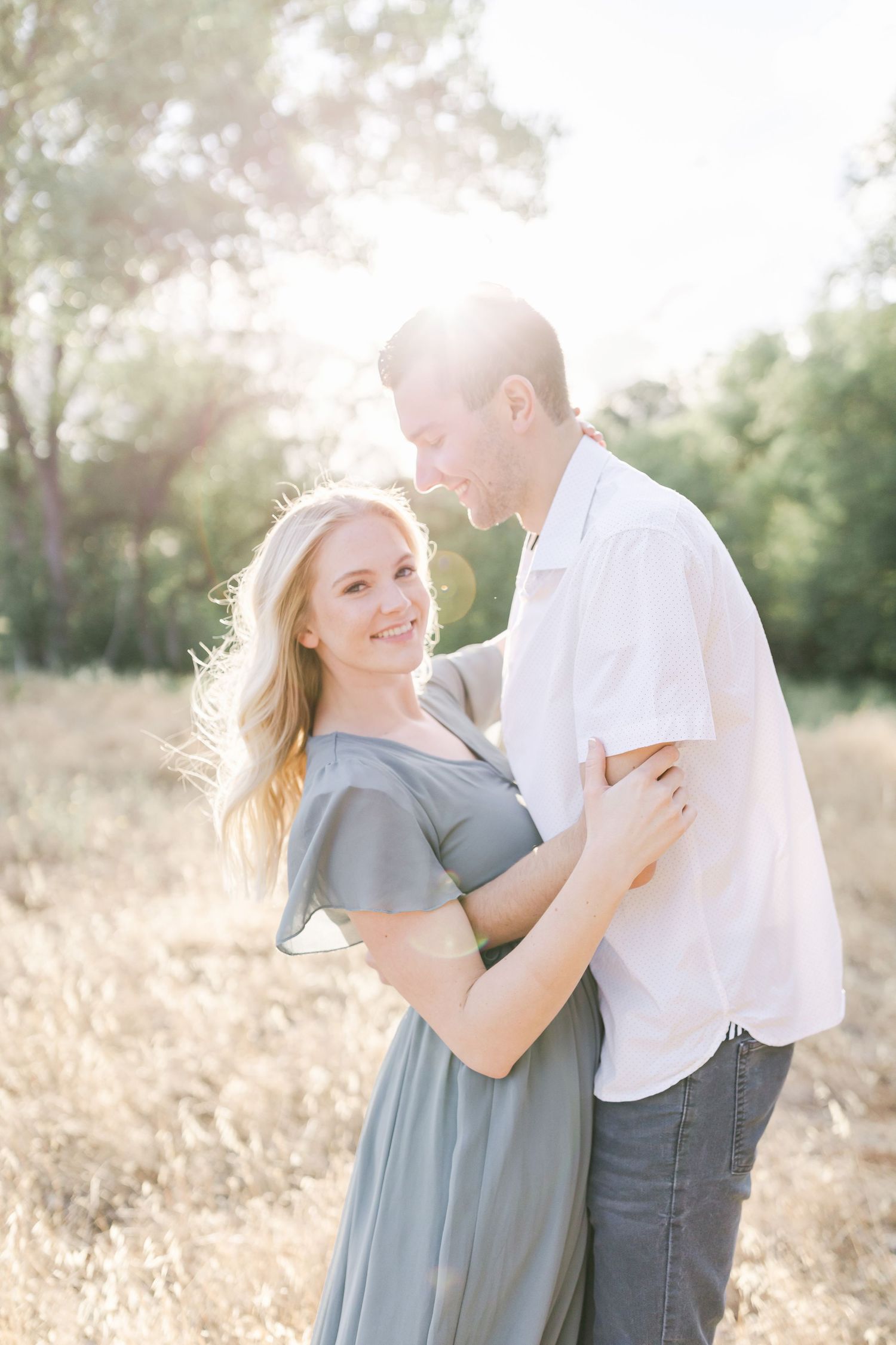 Loving couple embraces in a sunlit field during golden hour photoshoot wearing white and grey outfits.