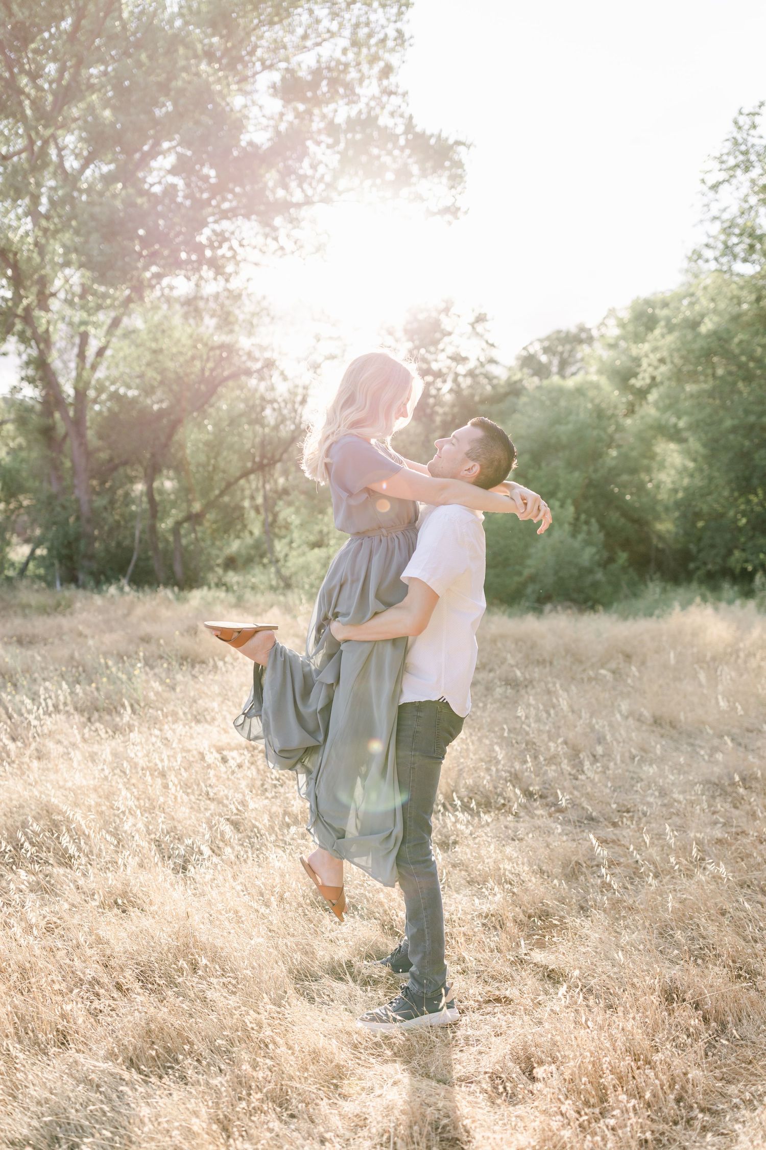 Romantic couple embracing in golden sunlit field during sunset with trees in background.
