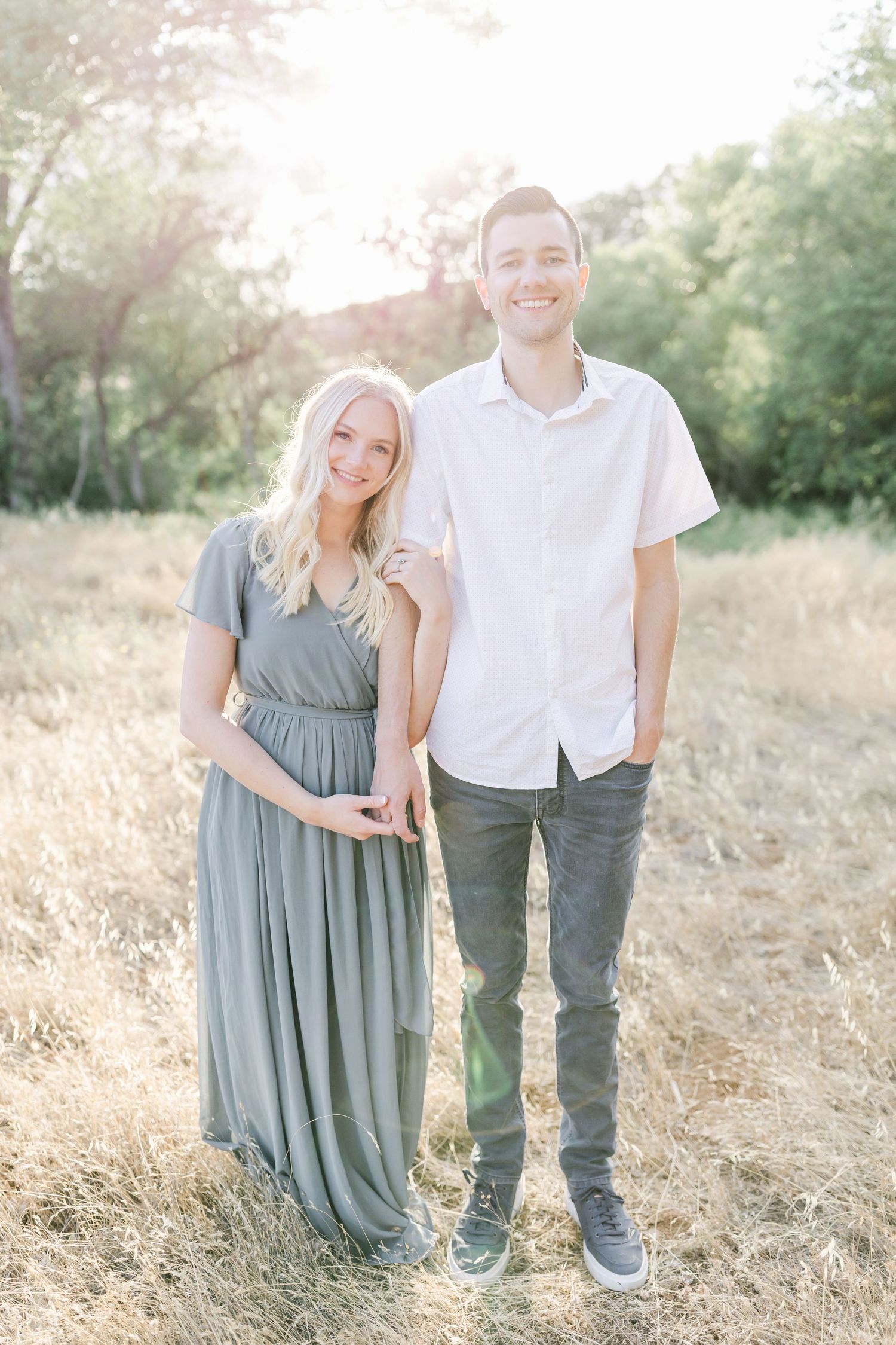 Couple standing together in a sunlit field wearing casual clothing and posing for engagement photos.