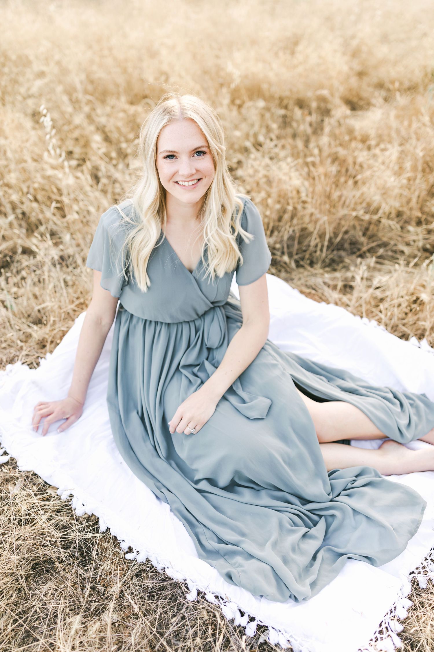 A flowy dusty blue dress elegantly draped on a white blanket in a golden wheat field on a sunny day Captured by Chelsee Rawe Photography