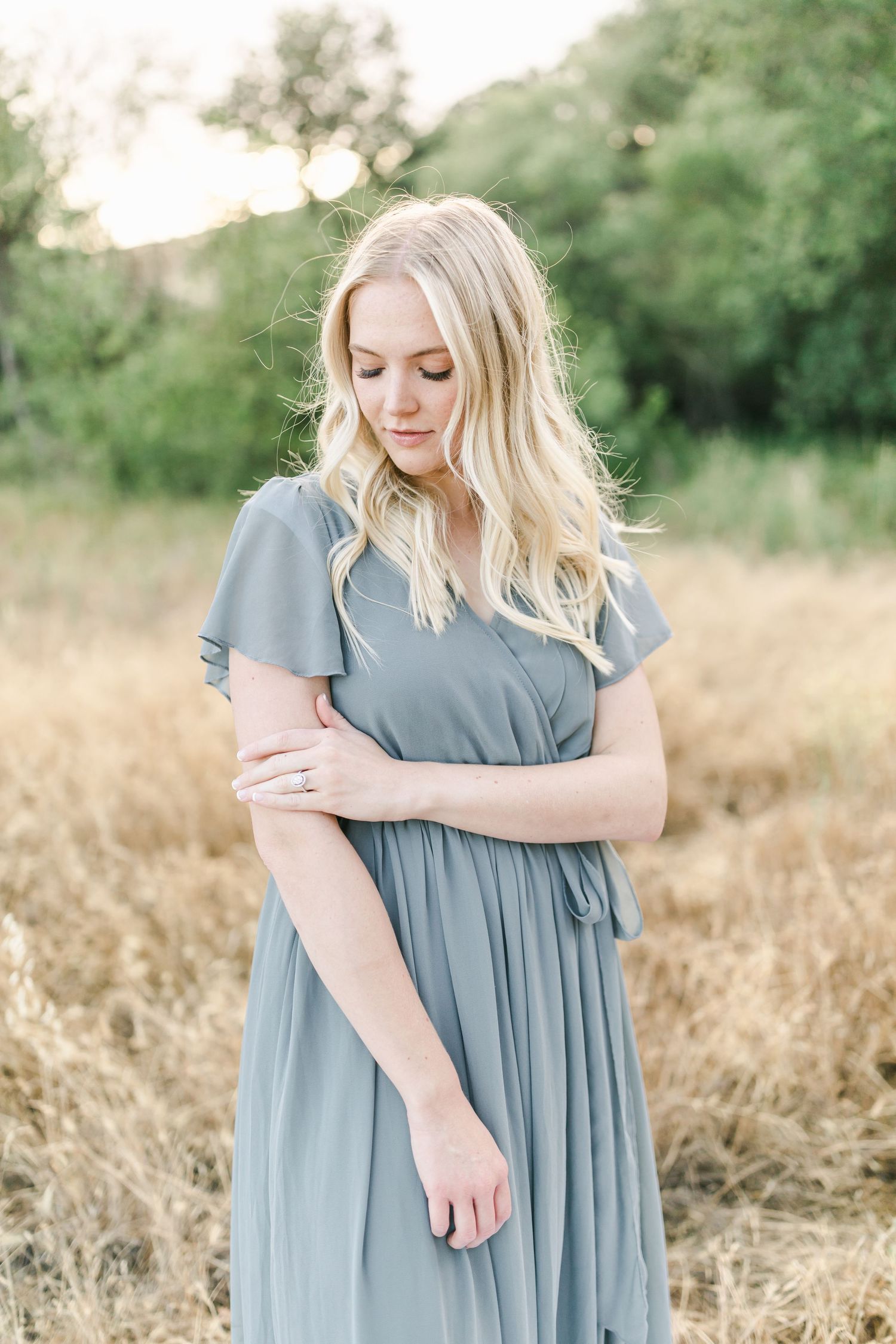 A woman in a flowing dusty blue dress stands thoughtfully in a golden wheat field at sunset.