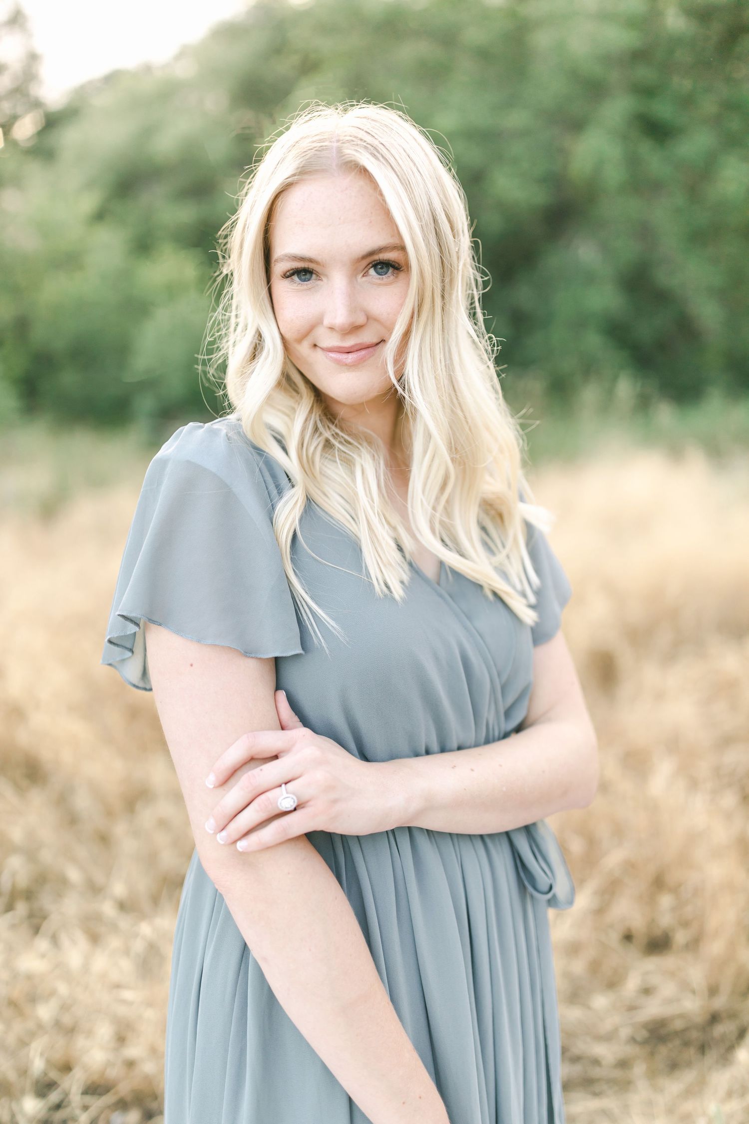 A person with blonde hair wearing a dusty blue flutter sleeve dress poses in a wheat field on a sunny day.