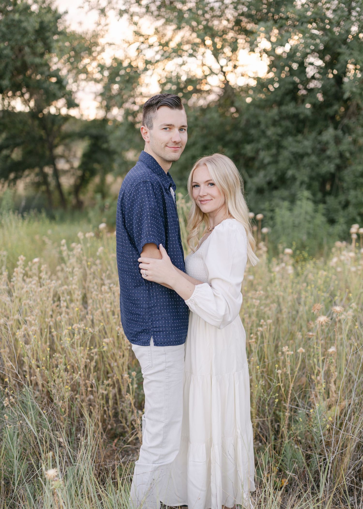 A couple embraces in a golden field at sunset with trees in the background.
