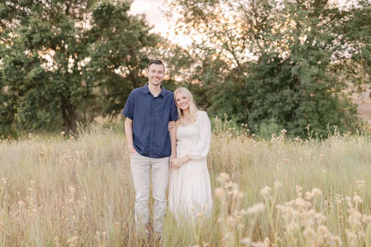 A couple stands together in a golden field at sunset with green trees in the background.
