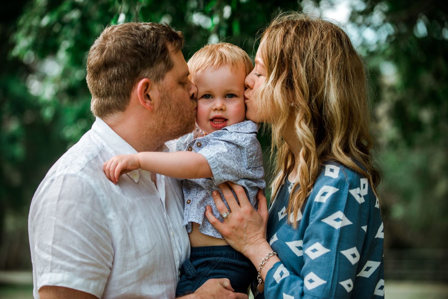 Young couple sharing tender moment with baby outdoors in natural setting with green foliage background.