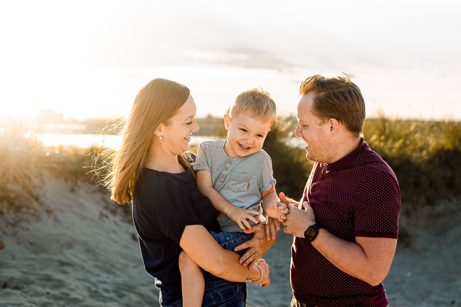 On a beach on Hayling Island, parents hold their three year old son close smiling and laughing in the sunlight.