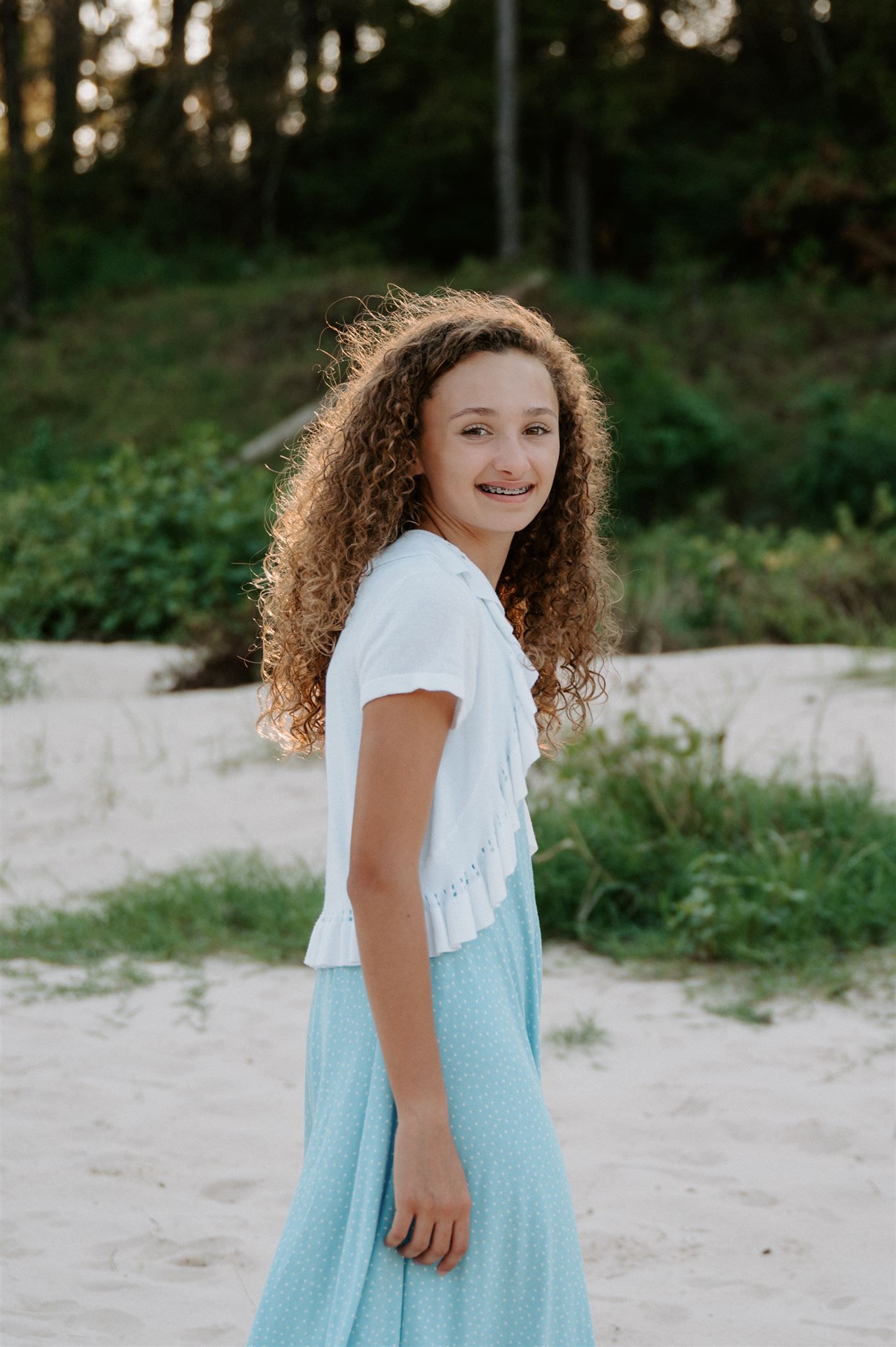 Young person with curly hair wearing blue skirt and white top smiles on sandy path.