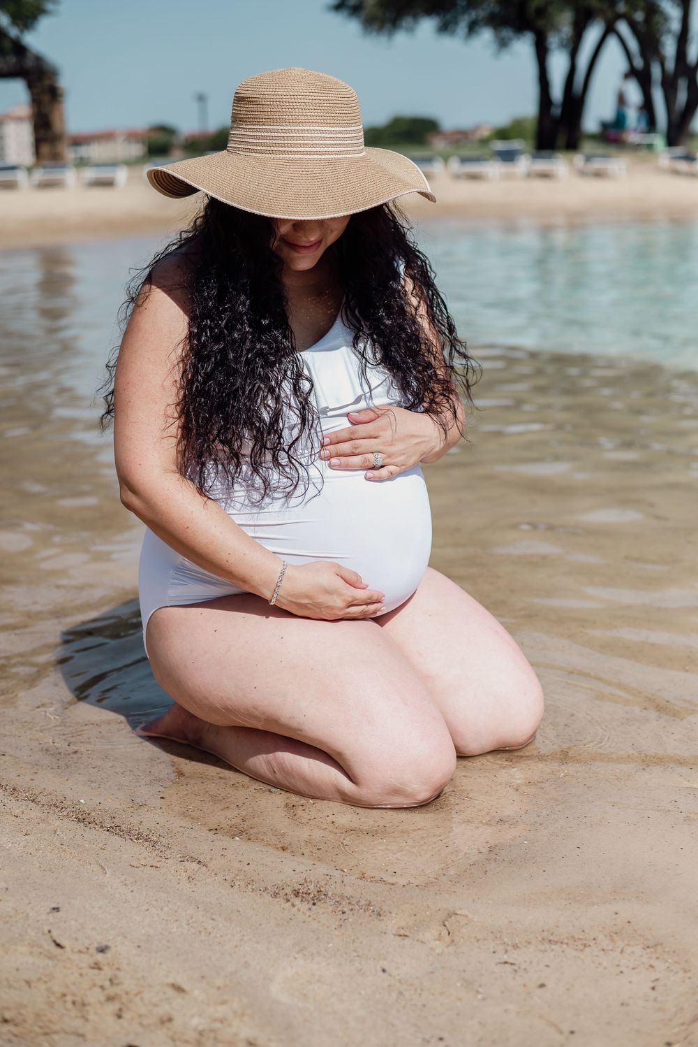 Person in white swimsuit and straw hat sitting on sandy beach near water.