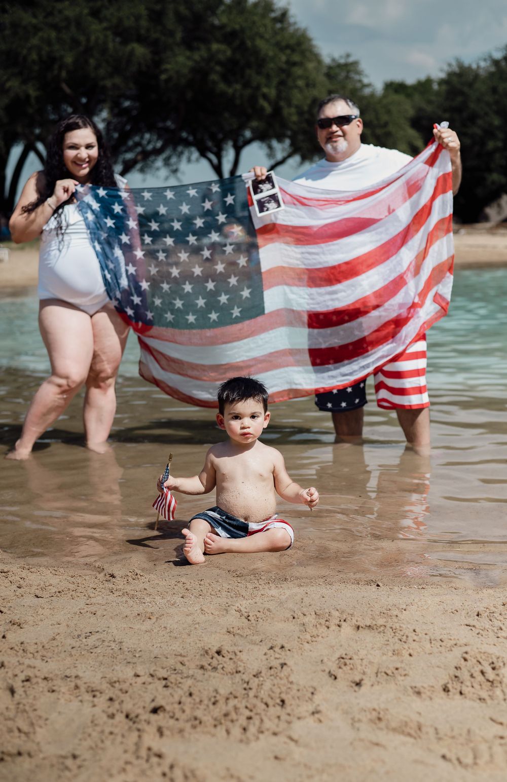 American flag held over sandy beach with small child sitting by water.