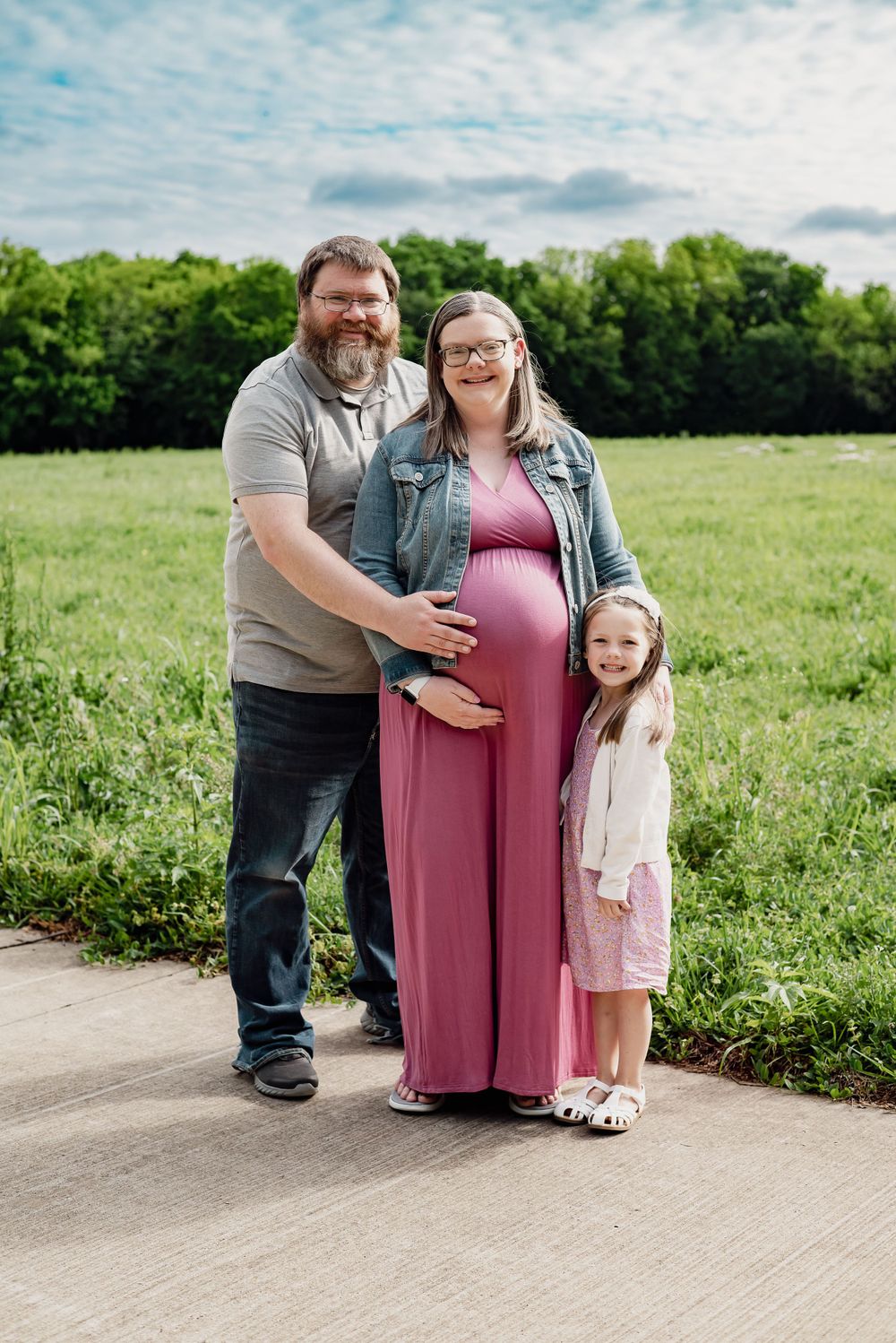 Family portrait outdoors on paved path with grassy field in background.
