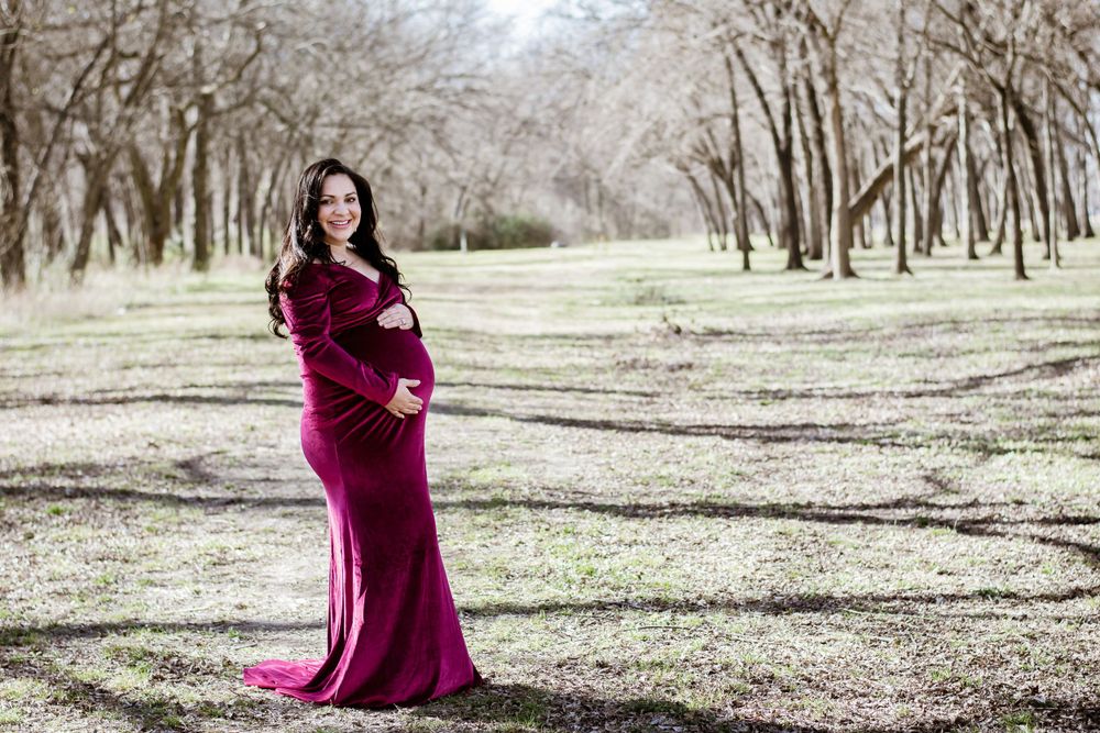 Woman in flowing burgundy velvet gown standing among winter trees.
