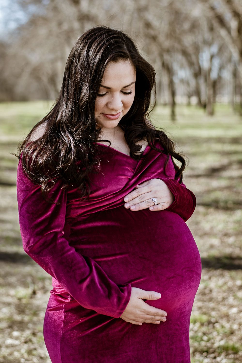 Woman in burgundy velvet dress cradling baby bump in wooded setting.