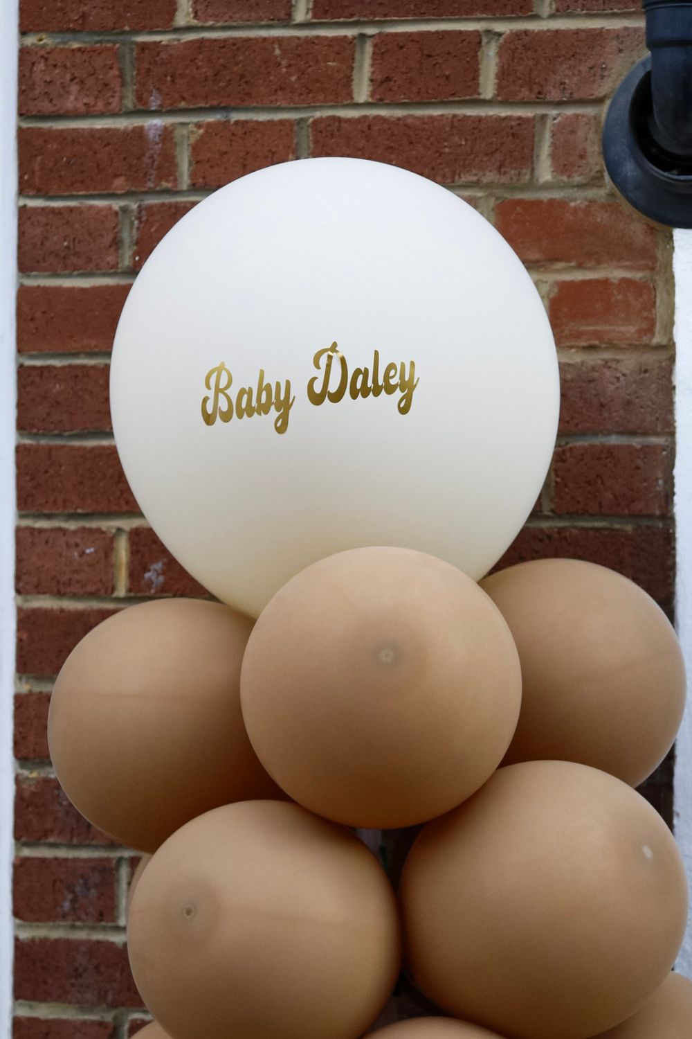 Gold lettering on white and brown balloon arrangement against brick wall background.