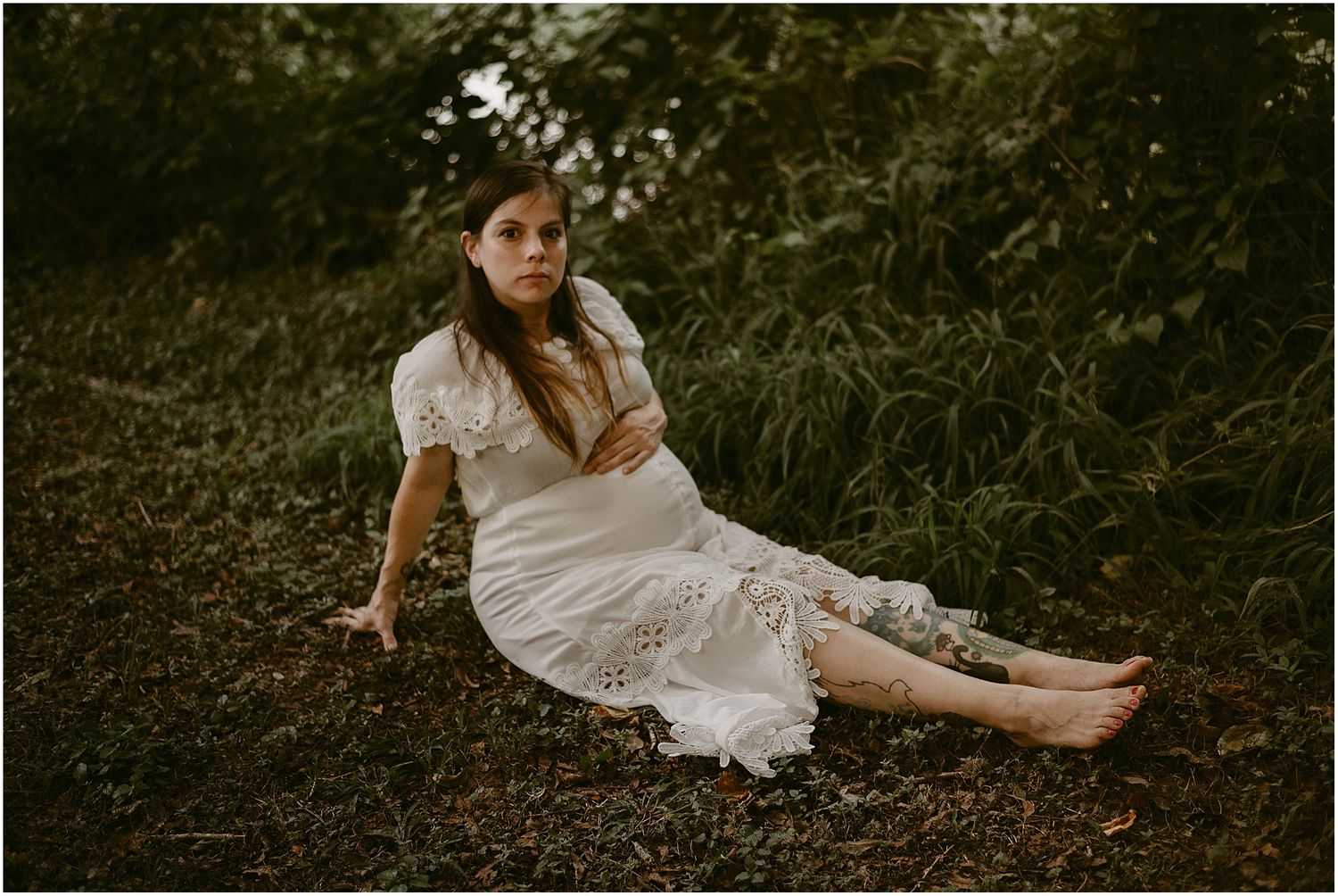Artistic maternity portrait on forest floor wearing vintage-inspired white lace dress.