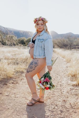 Maternity photoshoot in desert setting with denim jacket and floral bouquet against mountain backdrop.