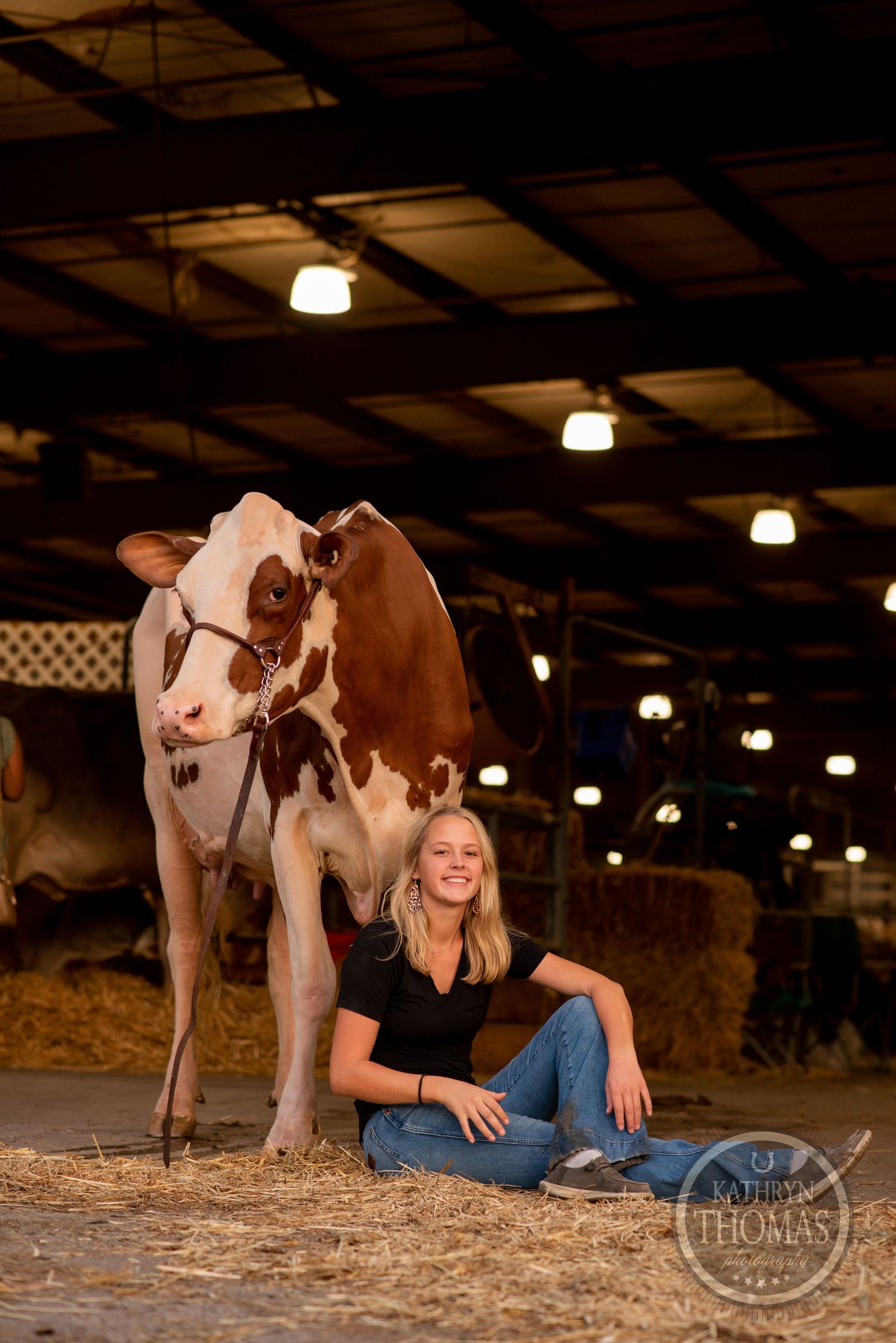 A dairy cow and person share a moment in a dimly lit barn with straw-covered floor.