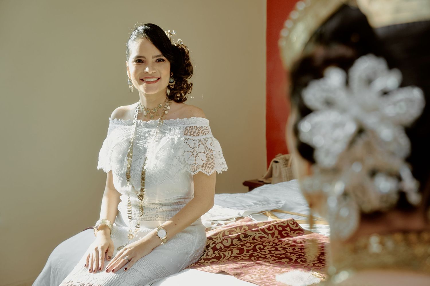 Elegant bride in off-shoulder lace wedding dress sitting gracefully with intricate headdress.