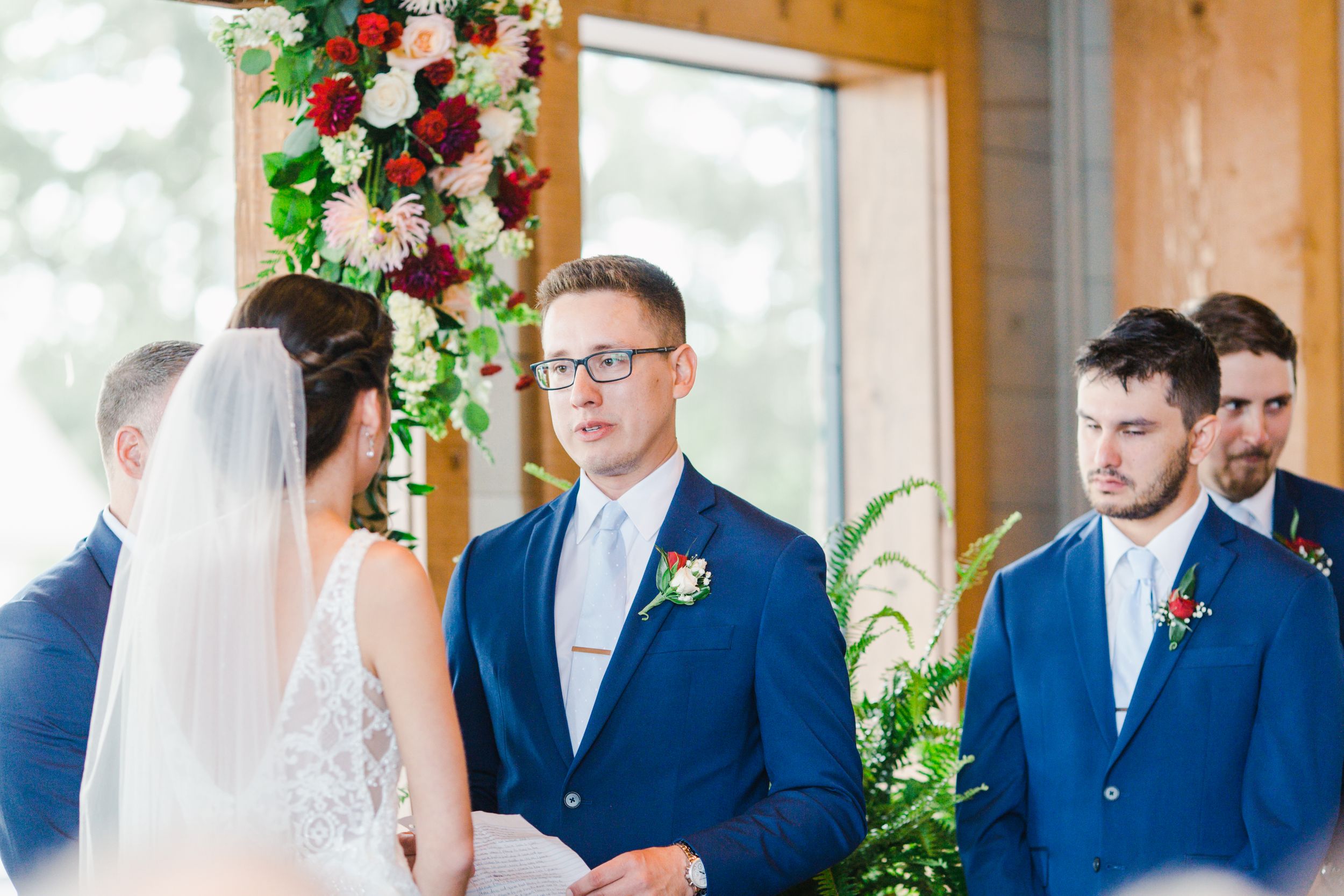 A wedding ceremony takes place in a sunlit venue decorated with red and white floral arrangements.
