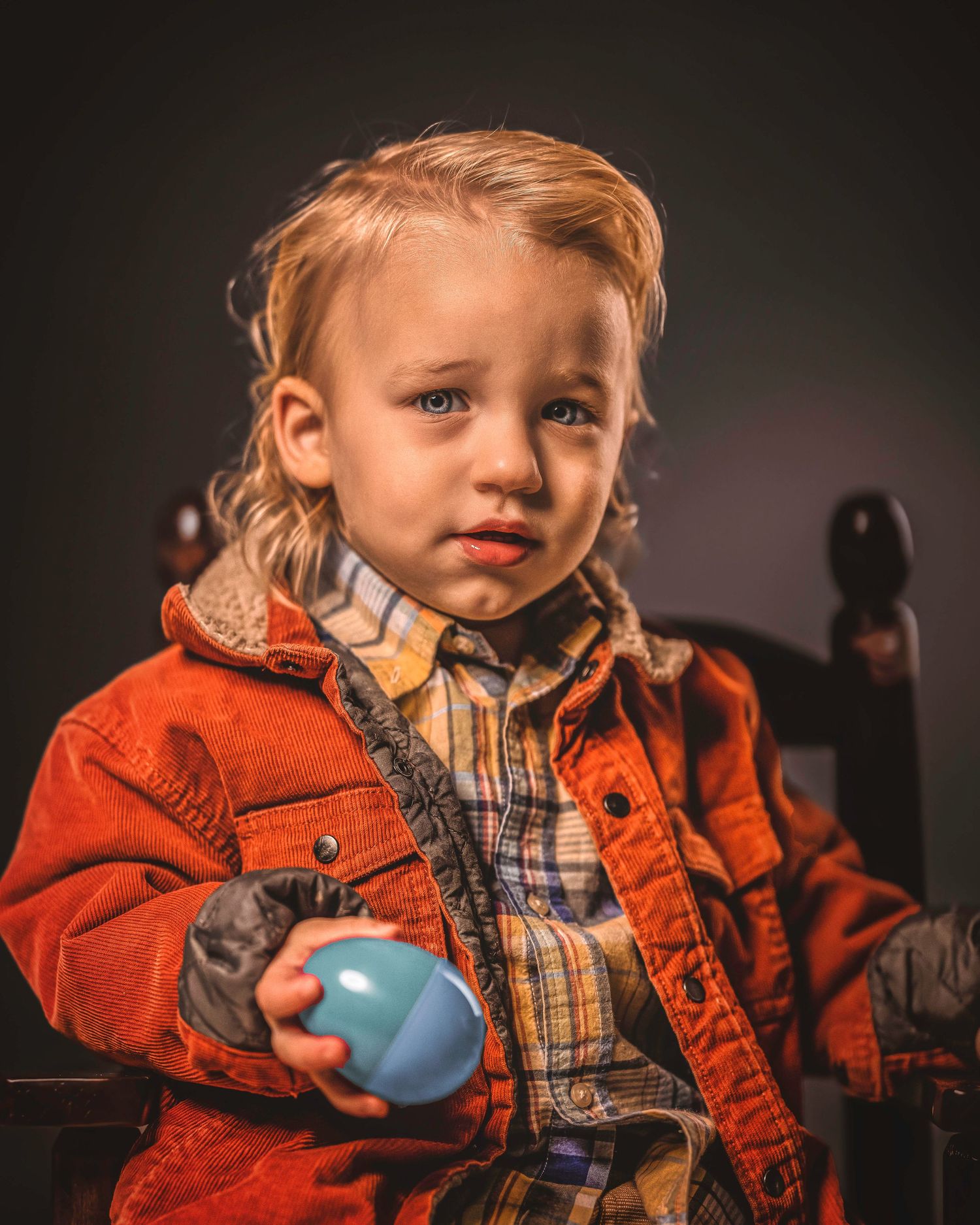 Adorable blonde toddler boy in an orange jacket holding a blue Easter egg in a studio setting.