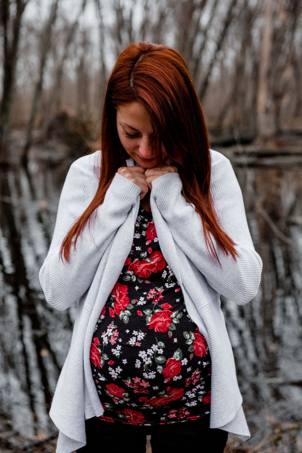 Woman in floral top and white cardigan poses near water in moody winter setting.