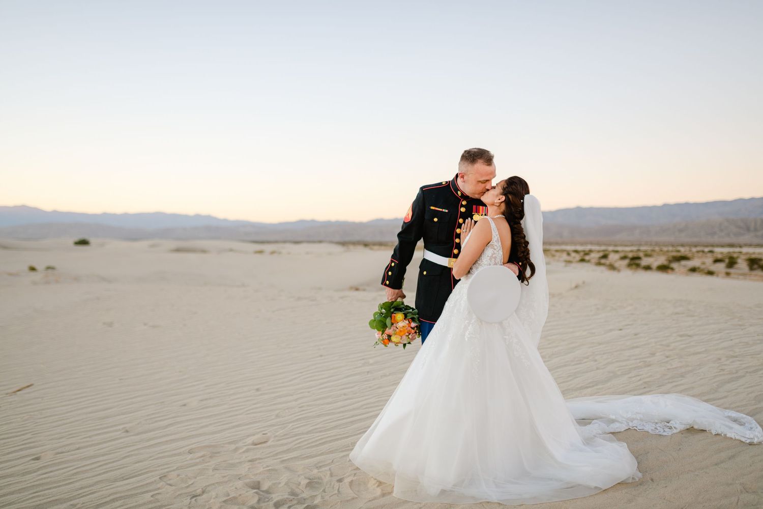A couple shares a romantic kiss in white wedding attire against a desert landscape backdrop.