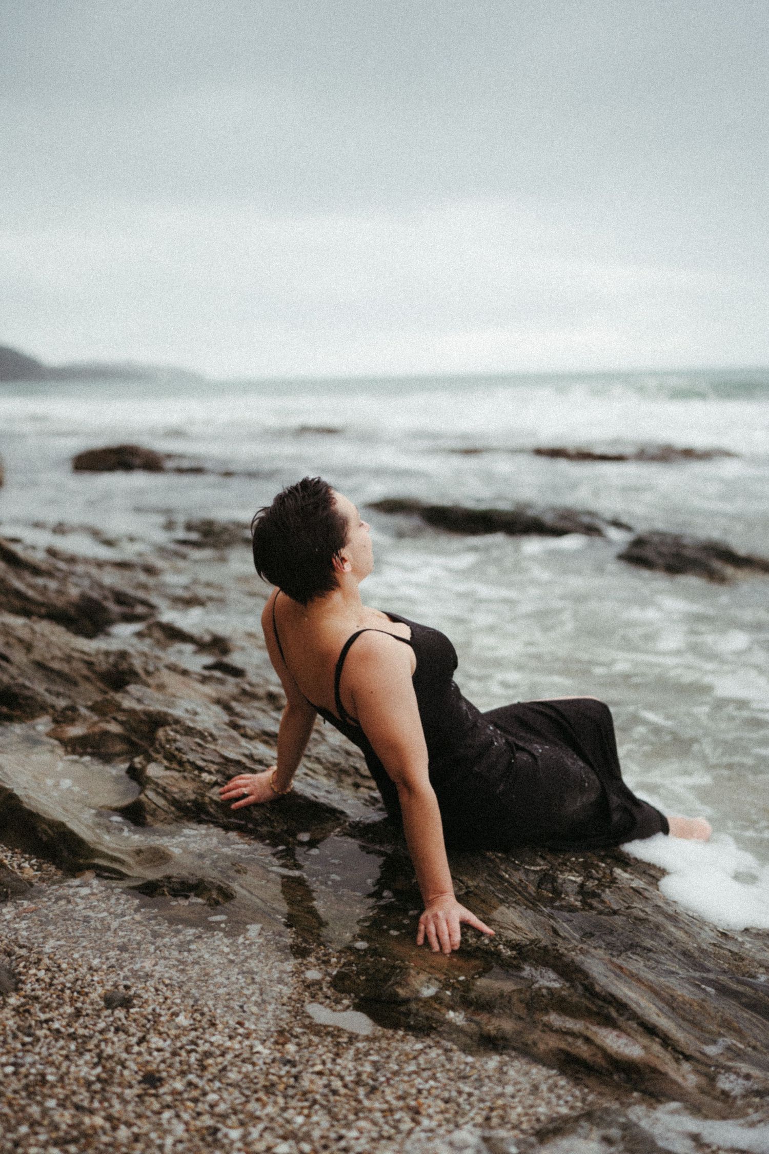 Waves crash against rocky shore as someone in black dress reclines on coastal rocks.