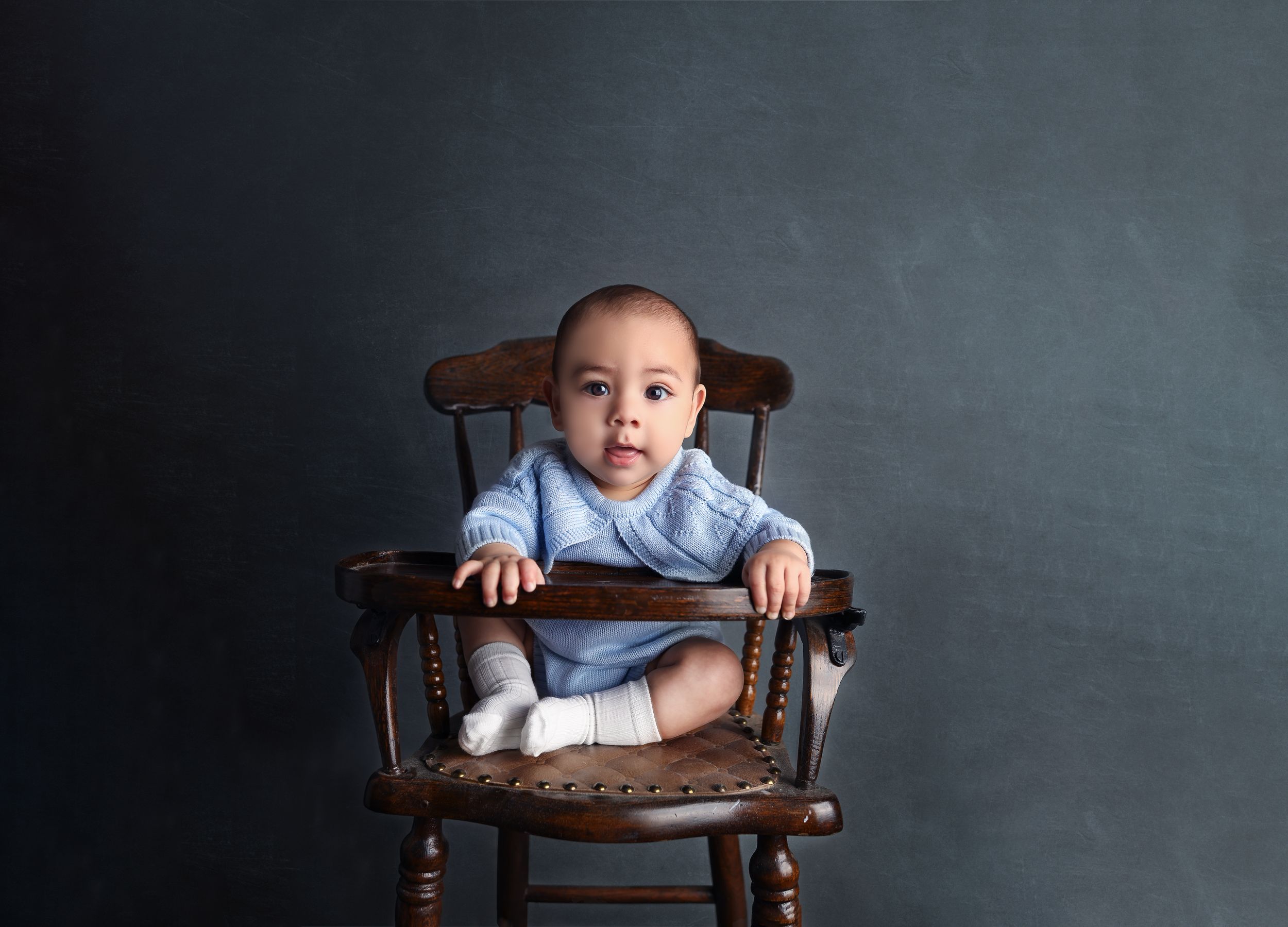 Baby dressed in blue sits in vintage wooden chair against grey backdrop.