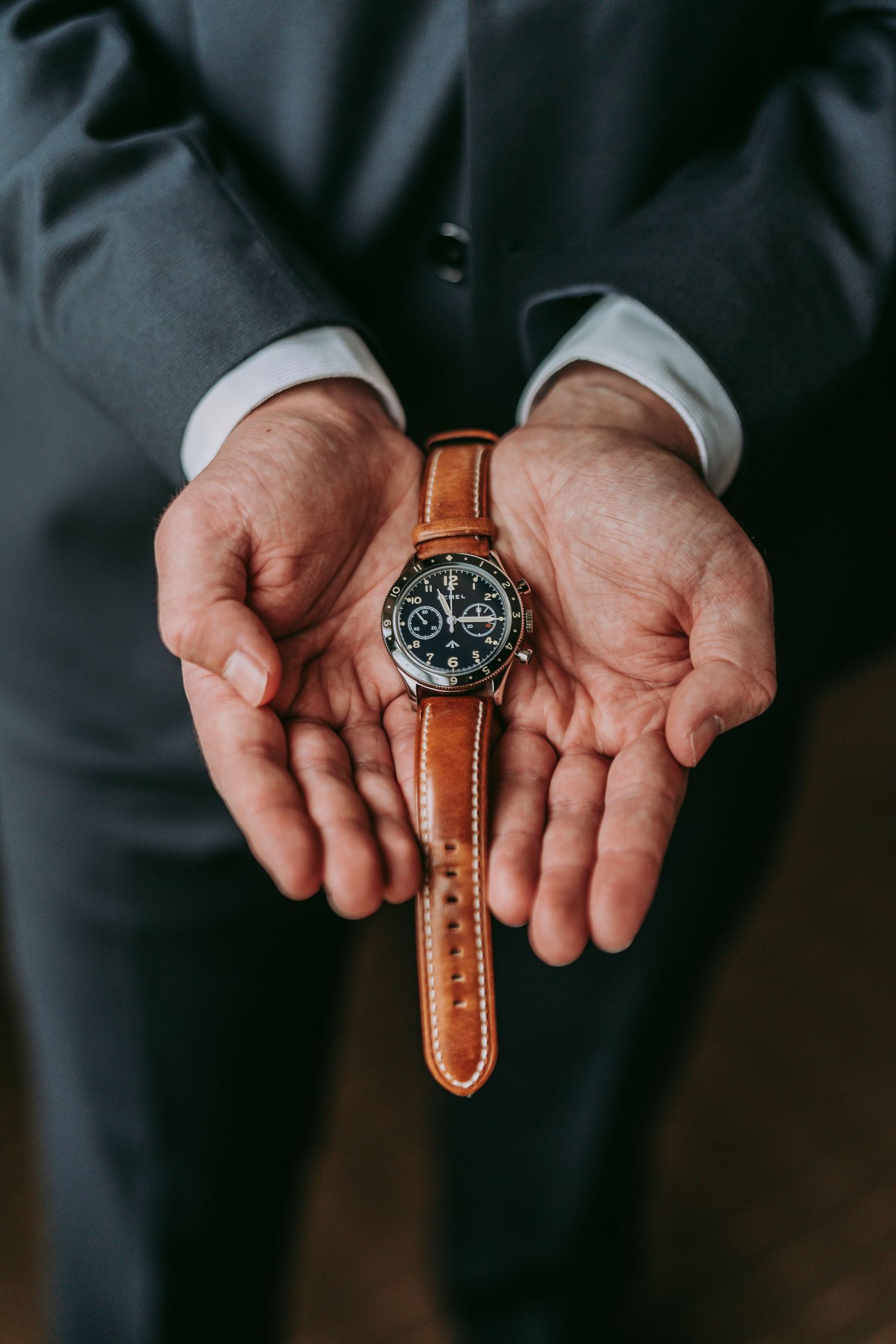 Hands holding a classic brown leather watch with chronograph face against dark suit.