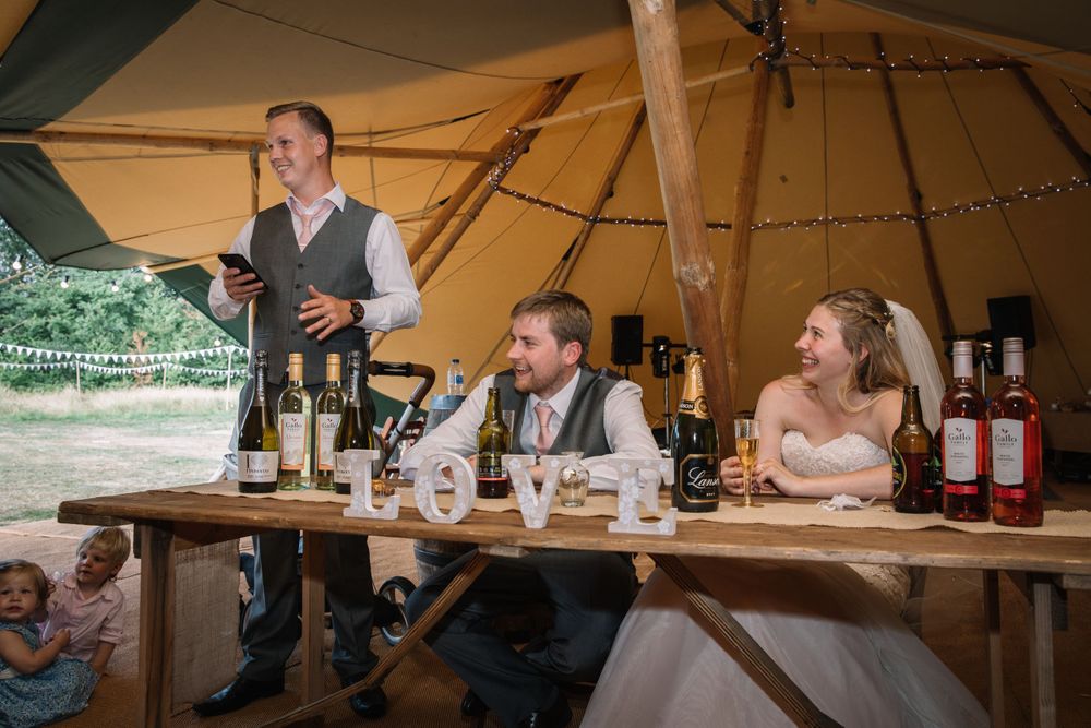 Wedding reception scene inside rustic tent with wooden tables and casual dining setup.