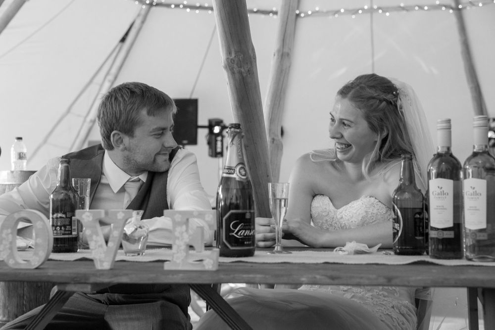 A black and white reception photo showing a couple seated at a dining table with wine bottles in a tent setting.