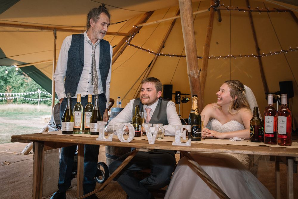Wedding reception guests gathered around a rustic wooden table inside a large canvas tent.