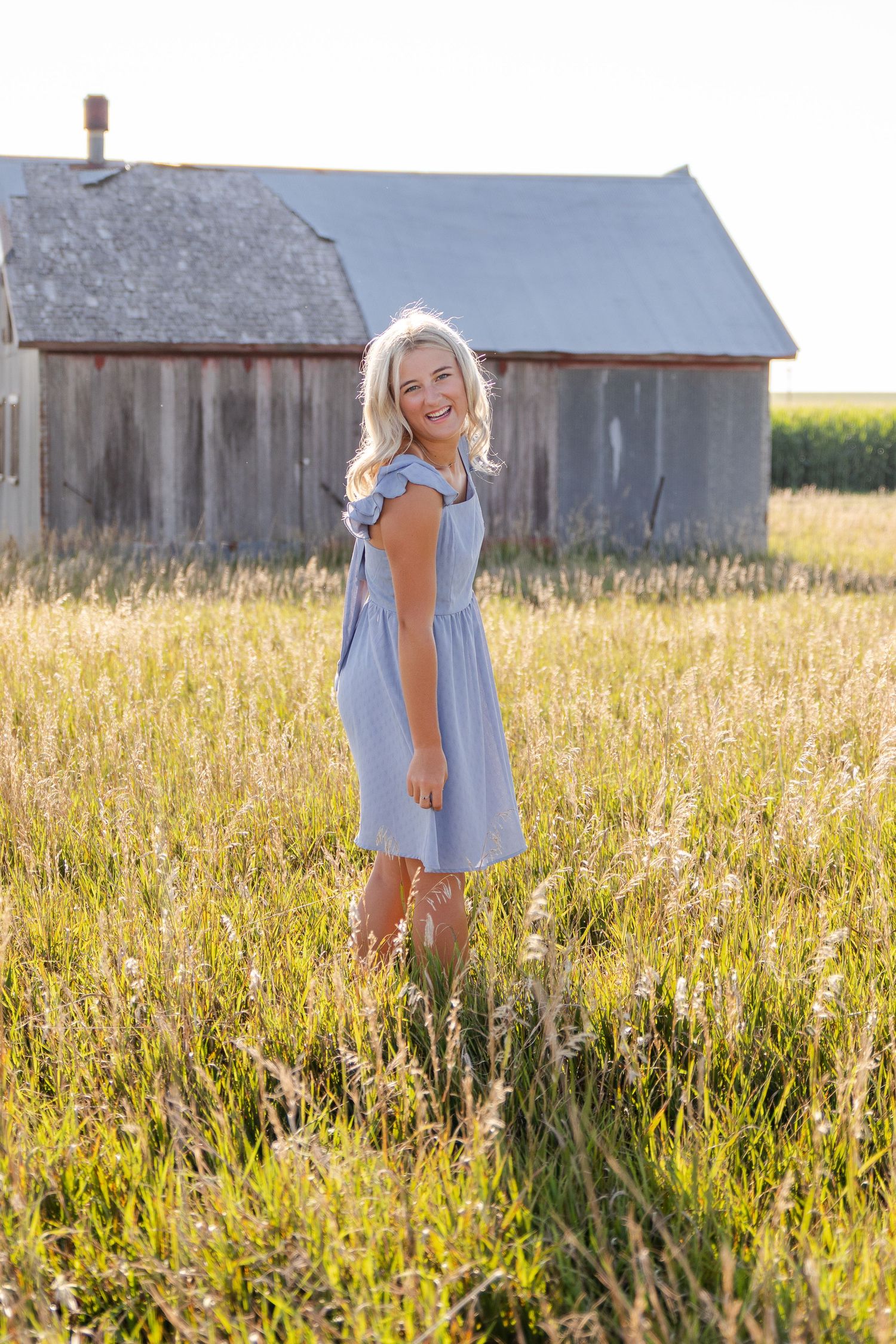 becky katzberg photography senior girl in blue dress standing in tall grass by metal barn