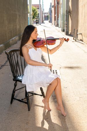becky katzberg photography senior girl playing violin in city alley wearing white dress and sitting on black chair