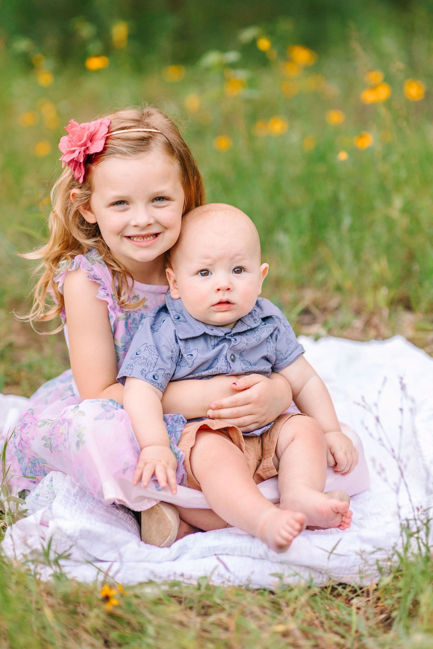 Sweet sibling moment captured on white blanket surrounded by yellow flowers.