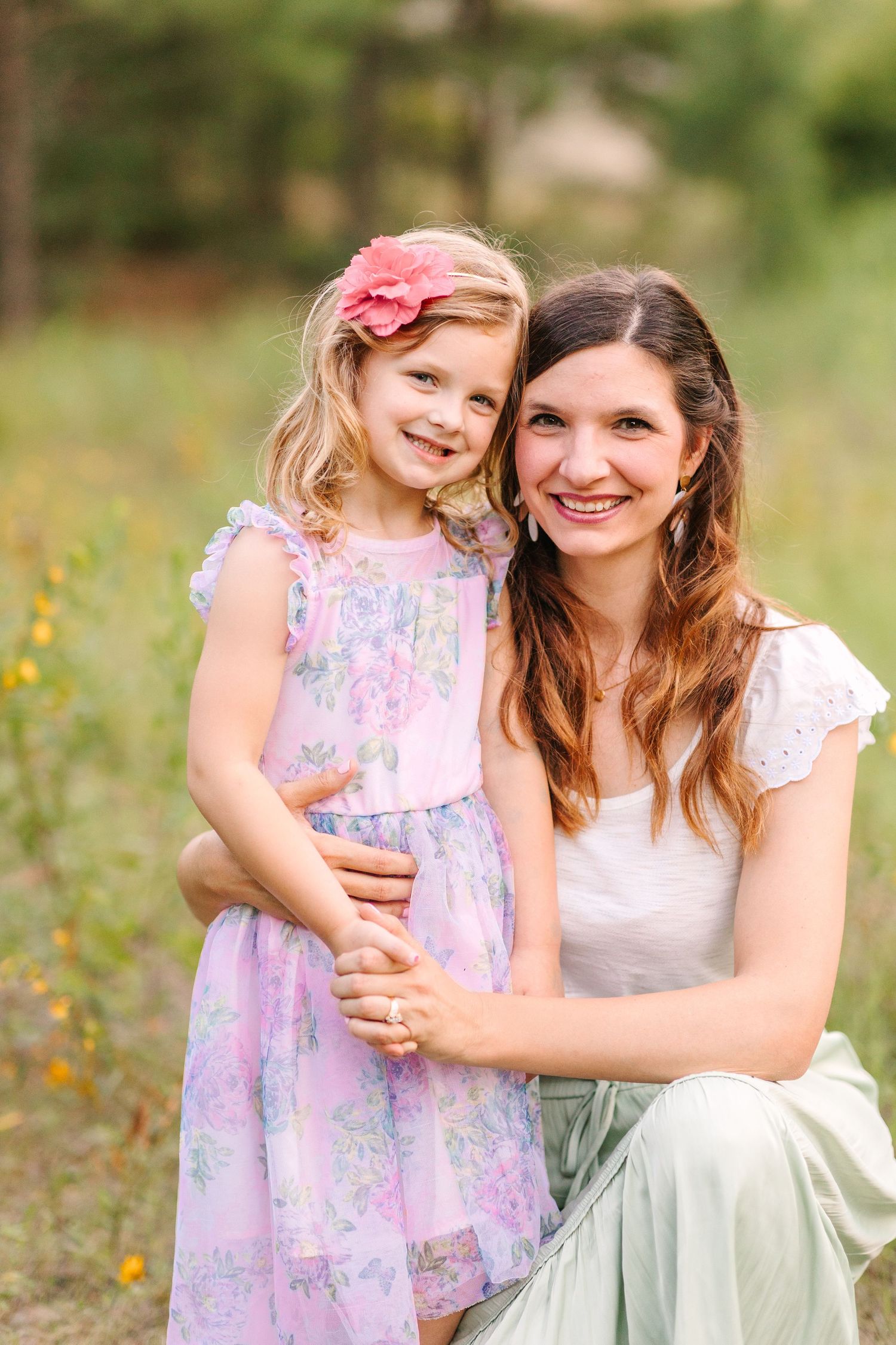 A mother and daughter in pastel dresses pose together in a sunny field of wildflowers.