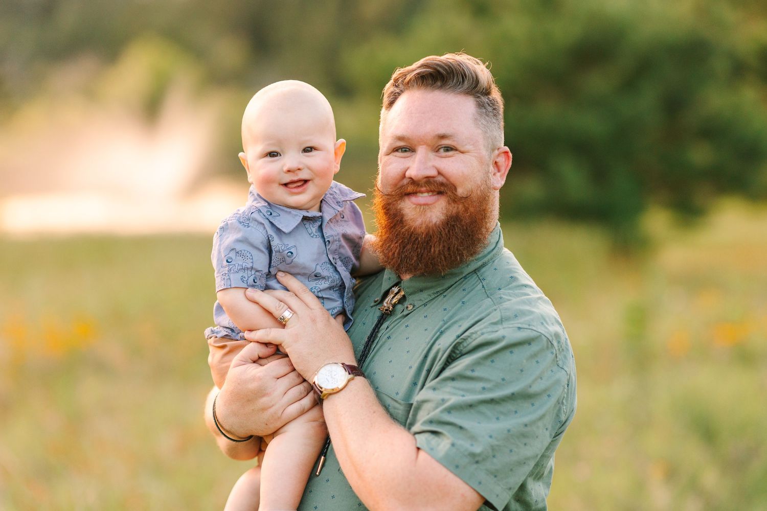 A bearded man in green shirt holds a smiling baby outdoors.