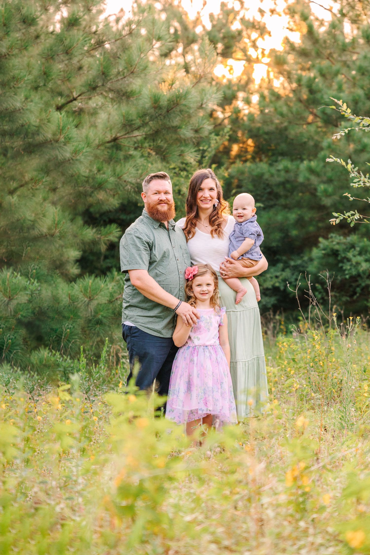 Family standing together in wildflower field during golden hour with pine trees in background.